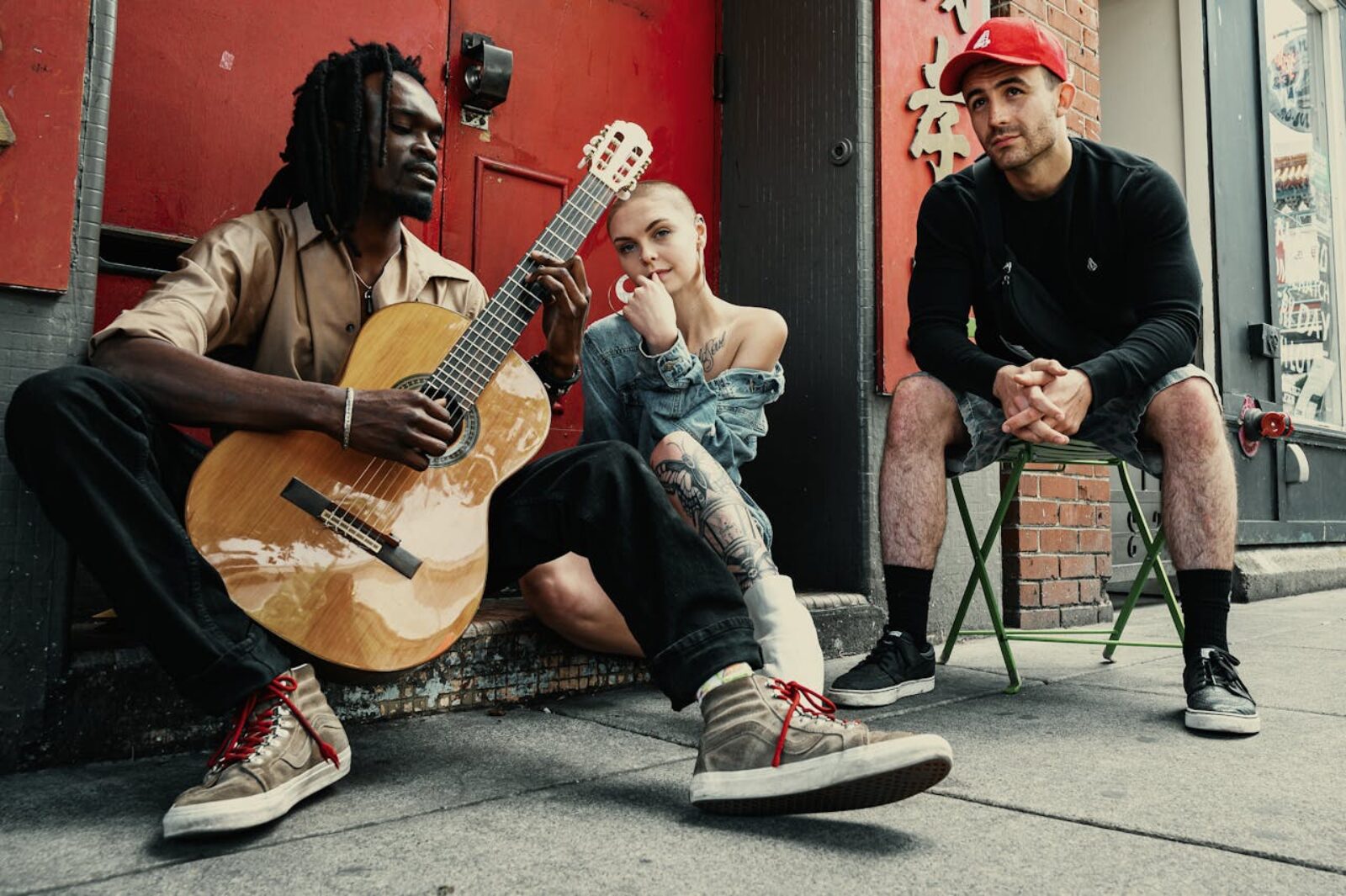Three diverse musicians sitting outdoors with guitar against red brick wall representing emerging artists seeking Rolling Stone feature