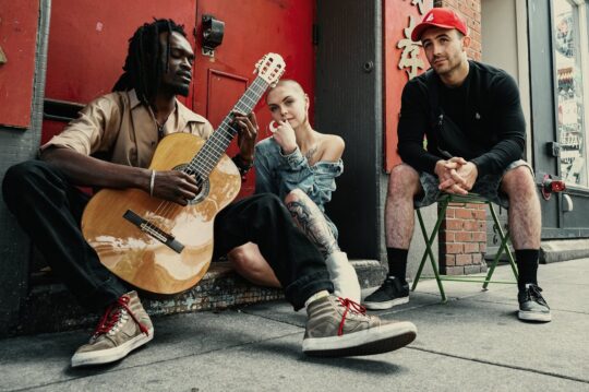 Three diverse musicians sitting outdoors with guitar against red brick wall representing emerging artists seeking Rolling Stone feature