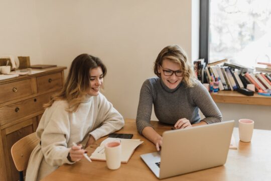 Two female digital marketers collaborating on technical SEO issues at wooden desk with laptop and notebooks