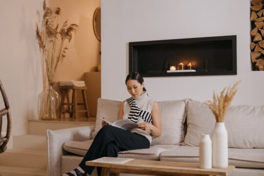 Professional woman reading Time Magazine while sitting on beige couch in modern living room with fireplace and candles