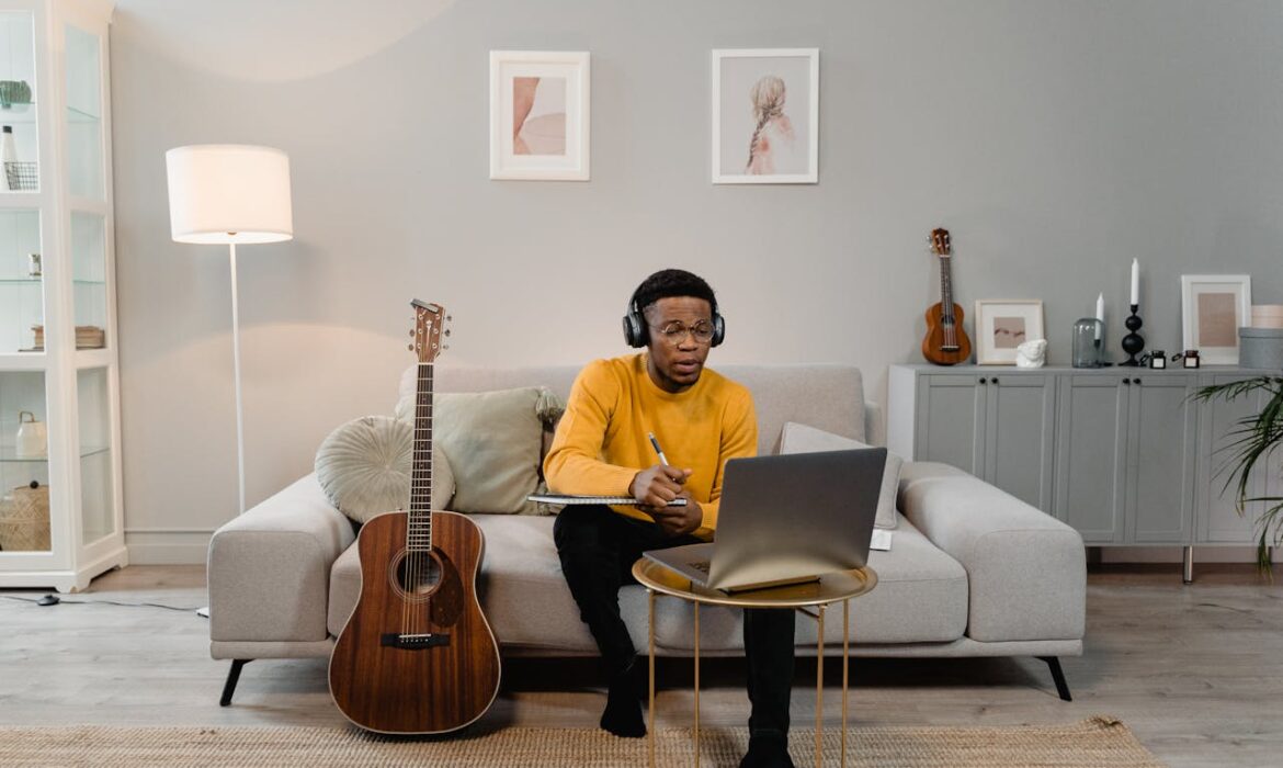 Independent musician wearing headphones taking notes on laptop in home studio with acoustic guitar