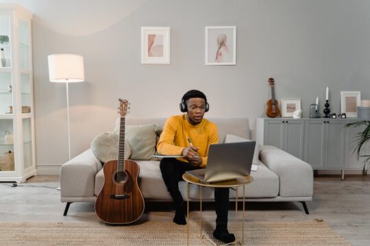 Independent musician wearing headphones taking notes on laptop in home studio with acoustic guitar