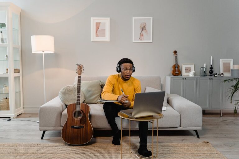 Independent musician wearing headphones taking notes on laptop in home studio with acoustic guitar